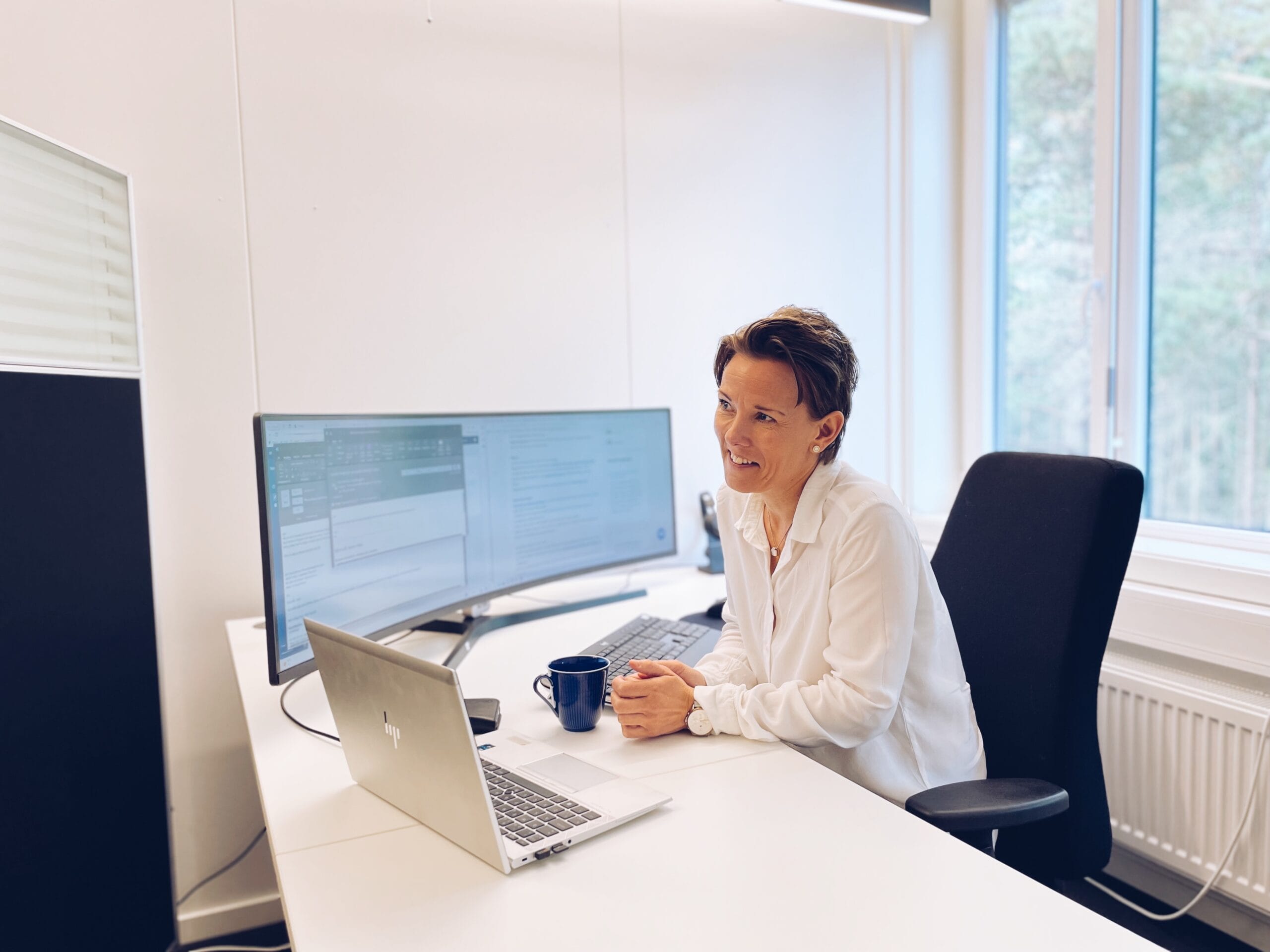 Kvinne med mørkt brunt hår i hvit skjorte sitter med sinn pult foran en stor skjerm og laptop.